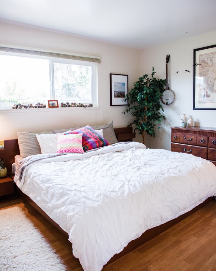 Cozy bedroom with a large bed, colorful pillows, wooden dresser, framed art, and a potted plant by the window.