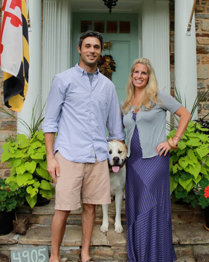 Couple with a dog standing on a stone porch, surrounded by plants, in front of a light blue door with a Maryland flag.