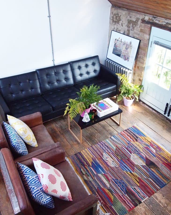 Loft living room with black leather sofa, colorful rug, wooden floor, and plants. Decor includes patterned cushions and framed art.
