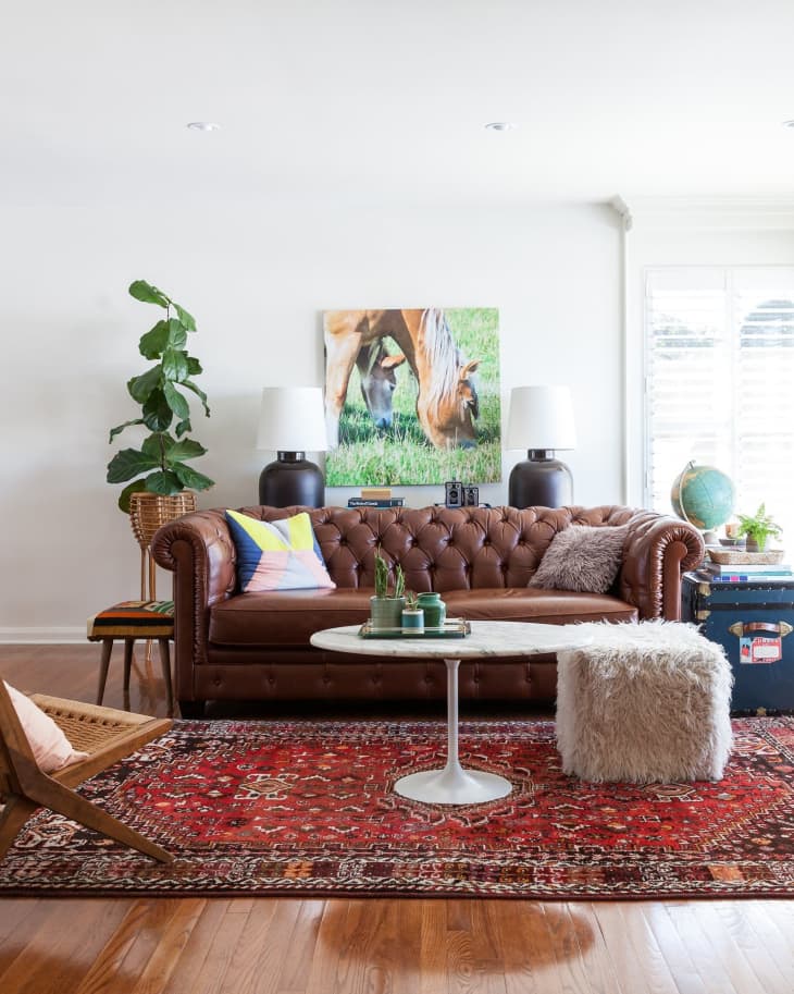 Brown leather sofa with colorful pillows, white round table, red patterned rug, and plant in a bright living room.