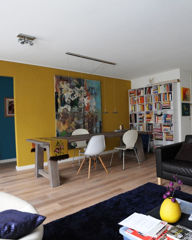 Modern living room with yellow accent wall, abstract art, wooden table, white chairs, bookshelves, and a black sofa.