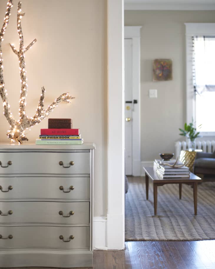Silver dresser with books and lit branch decor beside a living room with a coffee table and stack of books.