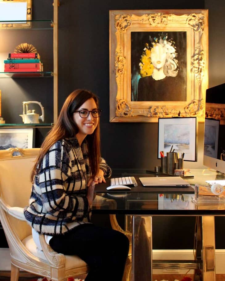 Woman in a plaid sweater sitting at a glass desk with a computer, framed art, and books on shelves in a dark room.