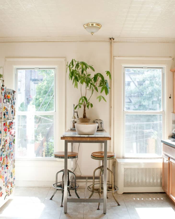 Bright kitchen with a tall wooden table, two stools, a potted plant, and a fridge covered in magnets.