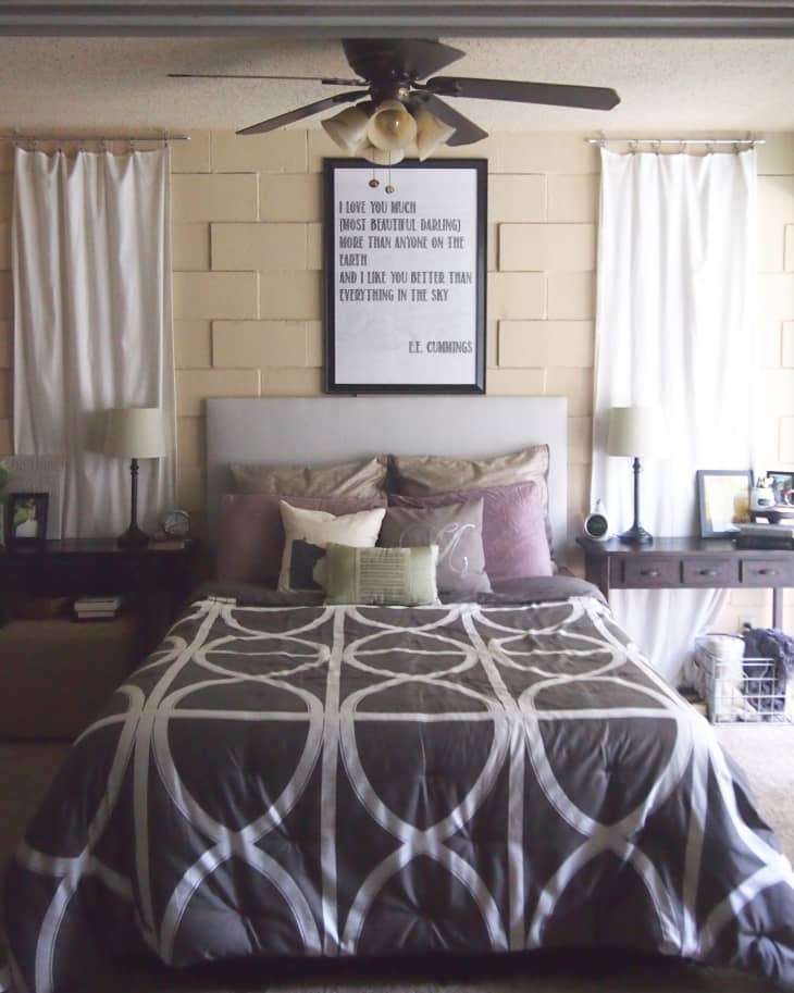 Bedroom with a patterned comforter, framed E.E. Cummings quote, ceiling fan, and bedside tables with lamps.