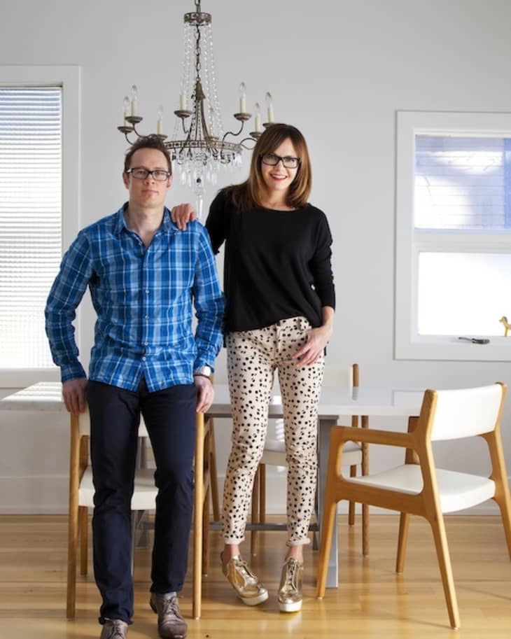 Two people in a bright dining room with wooden chairs and a chandelier.