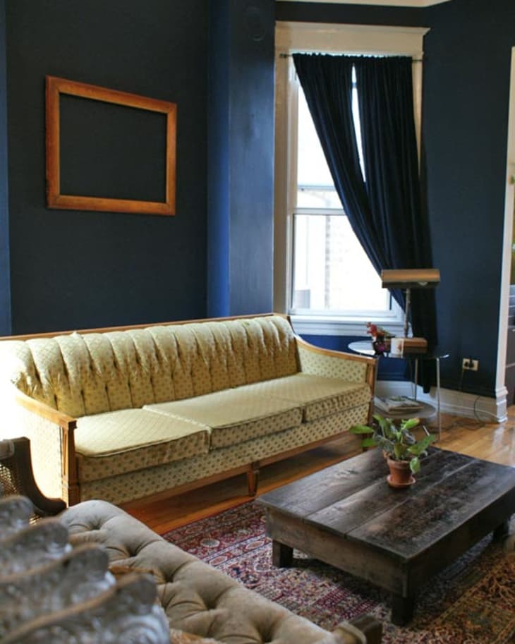 Vintage living room with a yellow sofa, dark wood coffee table, navy walls, and a window with dark curtains.
