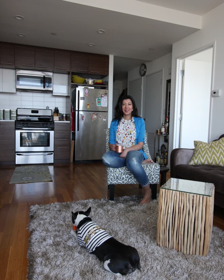 Woman sitting in a living room with a Boston Terrier on a rug, near a kitchen with stainless steel appliances.