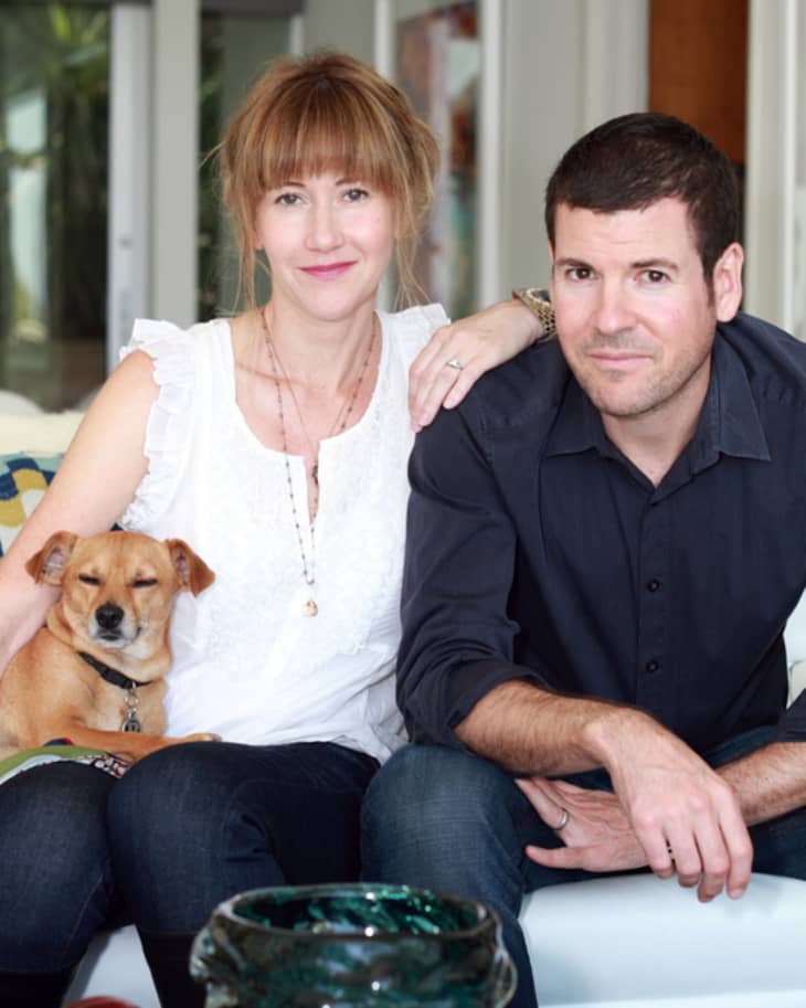 Couple sitting on a sofa with a small dog, surrounded by colorful glass decor in a modern living room.