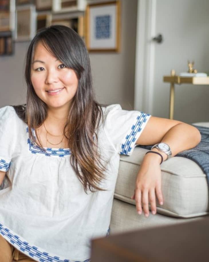 Woman in a white blouse with blue embroidery sitting on a beige sofa in a living room with framed art on the wall.