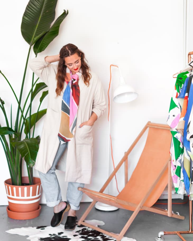 Woman in a beige coat and colorful scarf stands by a plant, orange deck chair, and clothes rack.