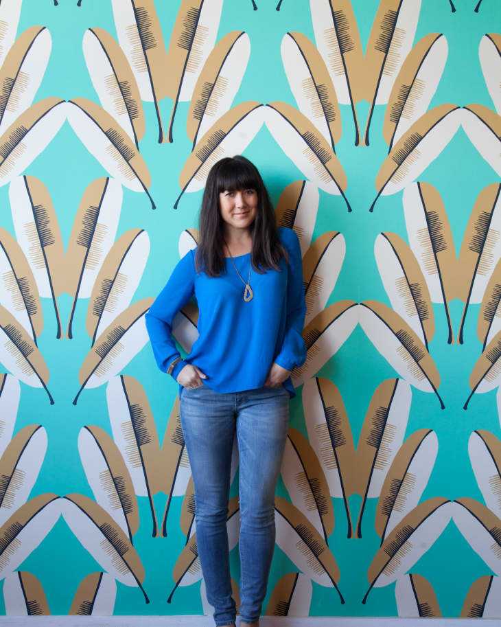 Woman in blue sweater and jeans standing against a teal feather-patterned wallpaper.