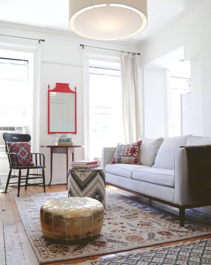 Living room with a gray sofa, patterned cushions, red mirror, wooden chair, gold pouf, and geometric rug.
