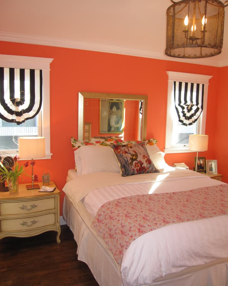 Bedroom with orange walls, floral pillows, striped curtains, and a vintage chandelier above a bed with a pink floral throw.