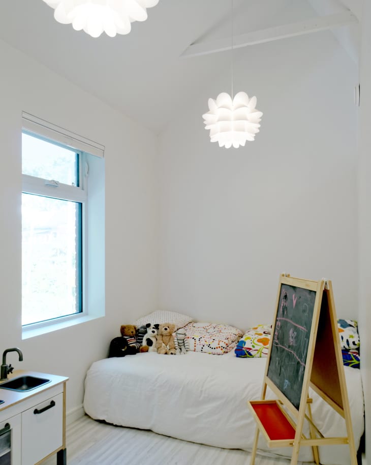 Children's room with a bed, colorful pillows, stuffed animals, a play kitchen, and a chalkboard easel under pendant lights.