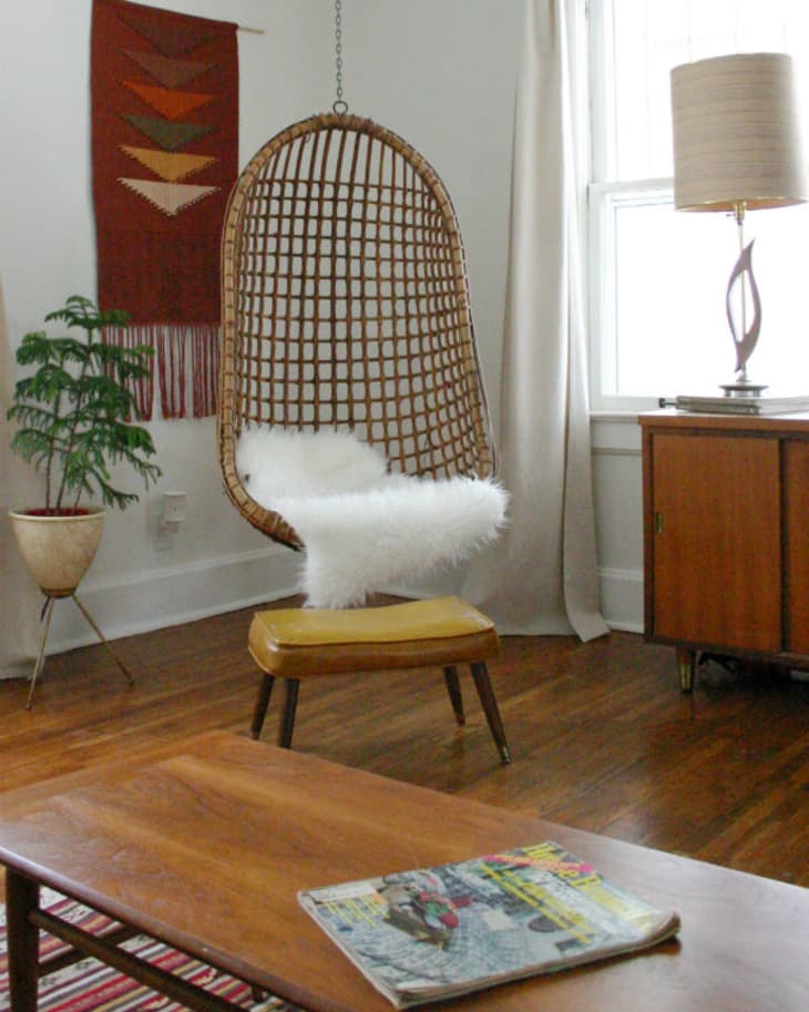 Hanging wicker chair with white cushion, wooden sideboard, potted plant, and red wall tapestry in a cozy living room.