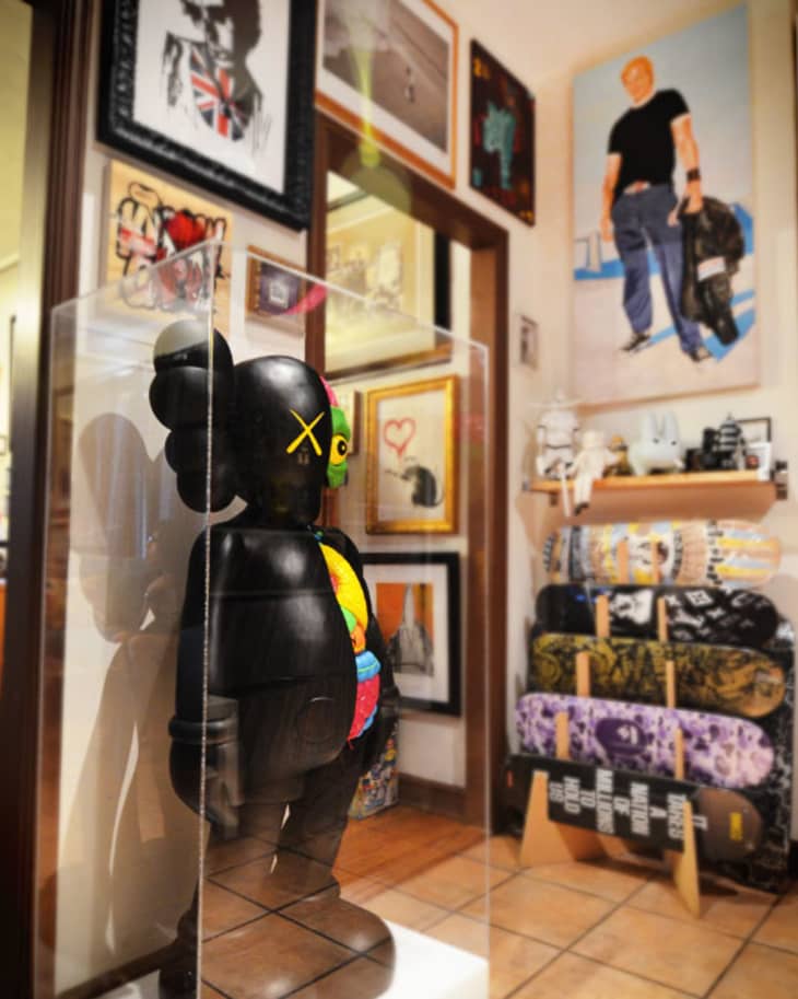 Art gallery with a black KAWS figure in a glass case, surrounded by framed artwork and stacked skateboards.