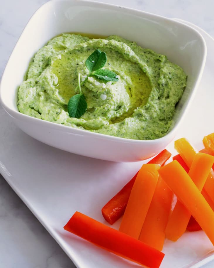 Creamy green dip garnished with a mint leaf, served with orange and red bell pepper sticks on a white plate.