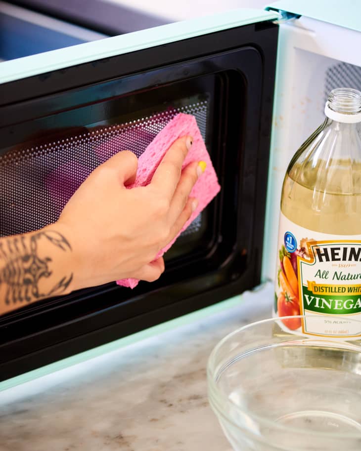 A person cleaning the inside of a microwave with a sponge and white vinegar