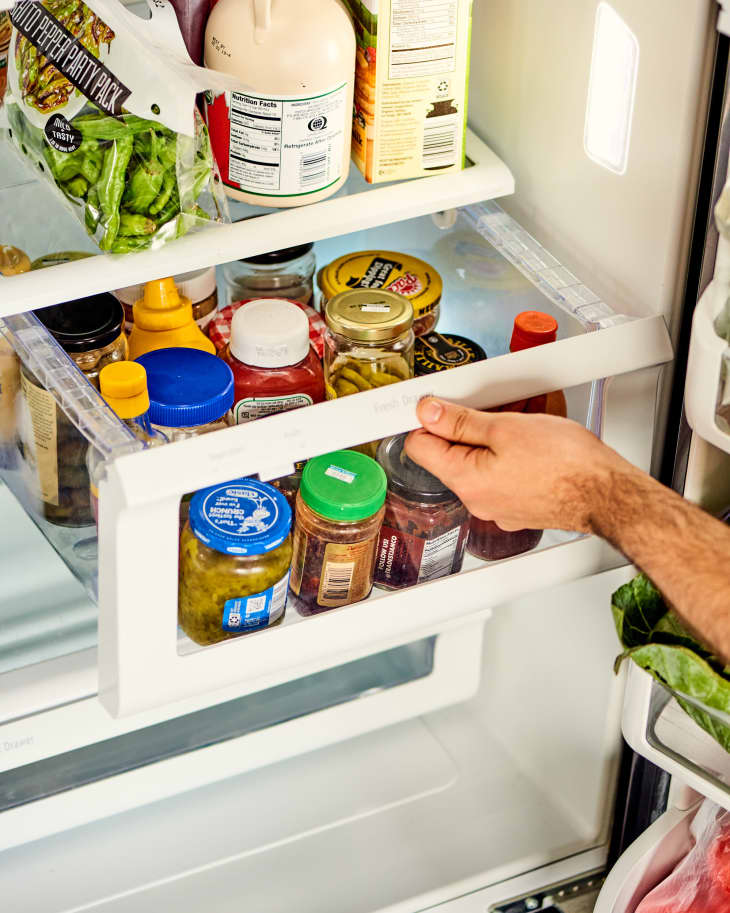 Hand pulling out a drawer/bin in a refrigerator. The bin is full of condiments (catsup, mustard, hot sauce, etc)