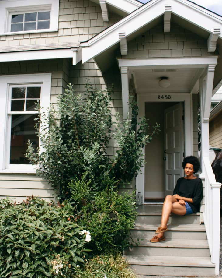 Woman sitting on steps of a gray house with white trim, surrounded by lush greenery and flowers.