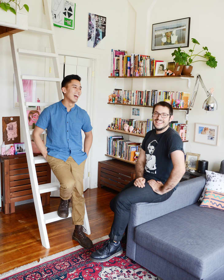 Two people in a cozy living room with bookshelves, a gray sofa, colorful rug, and wall art.