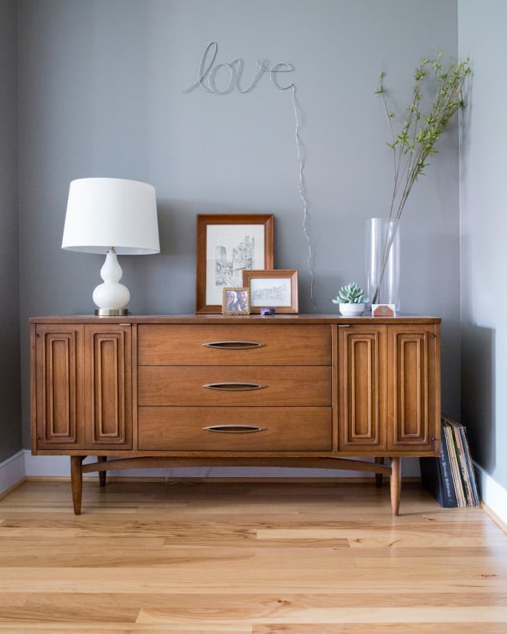 Mid-century modern wooden sideboard with framed art, a white lamp, and a vase with branches, under a "love" wire sign.