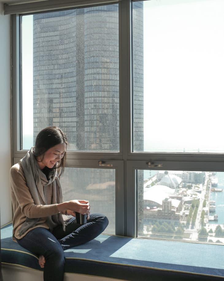 Woman sitting on a window seat, holding a mug, with a cityscape view outside.