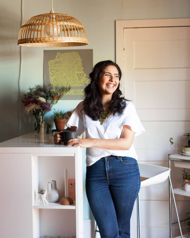 Woman in white shirt and jeans holding a mug, standing by a kitchen counter with a wicker pendant light above.
