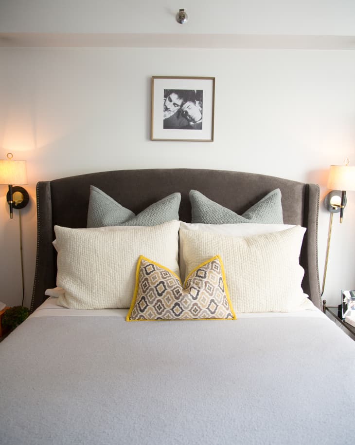 Gray upholstered bed with white and patterned pillows, flanked by wall sconces, and a framed black-and-white photo above.