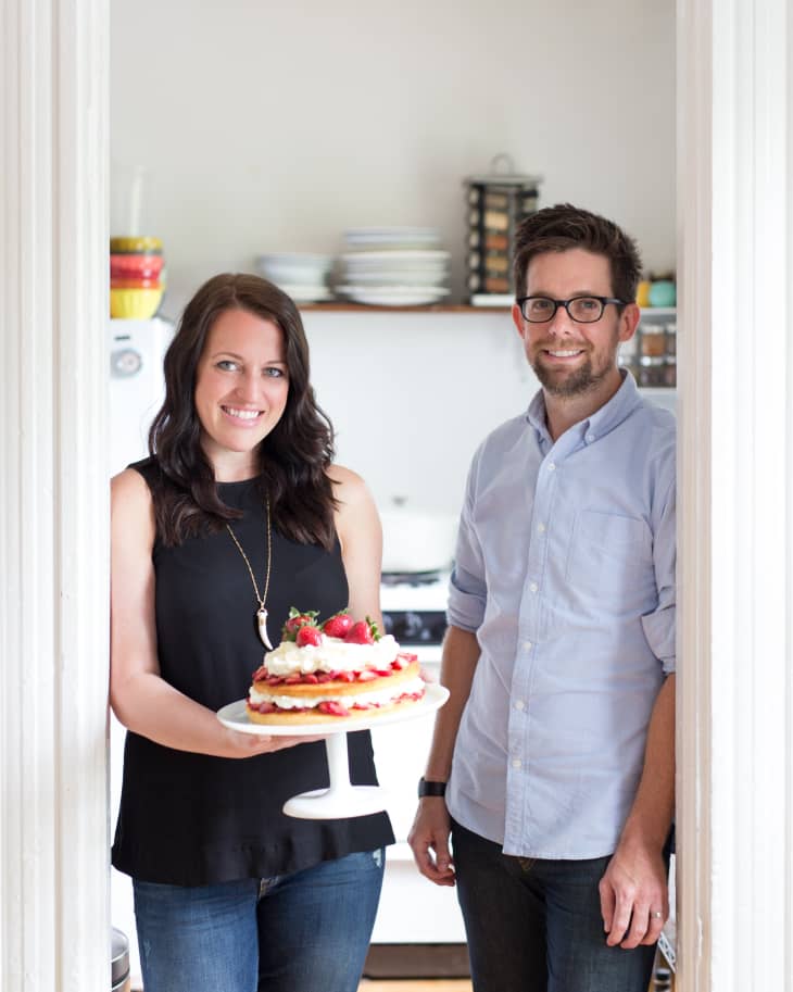Woman holding a strawberry-topped cake on a stand, standing next to a man in a kitchen doorway.