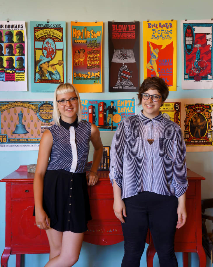 Two people standing in front of a red table with colorful vintage posters on the wall.