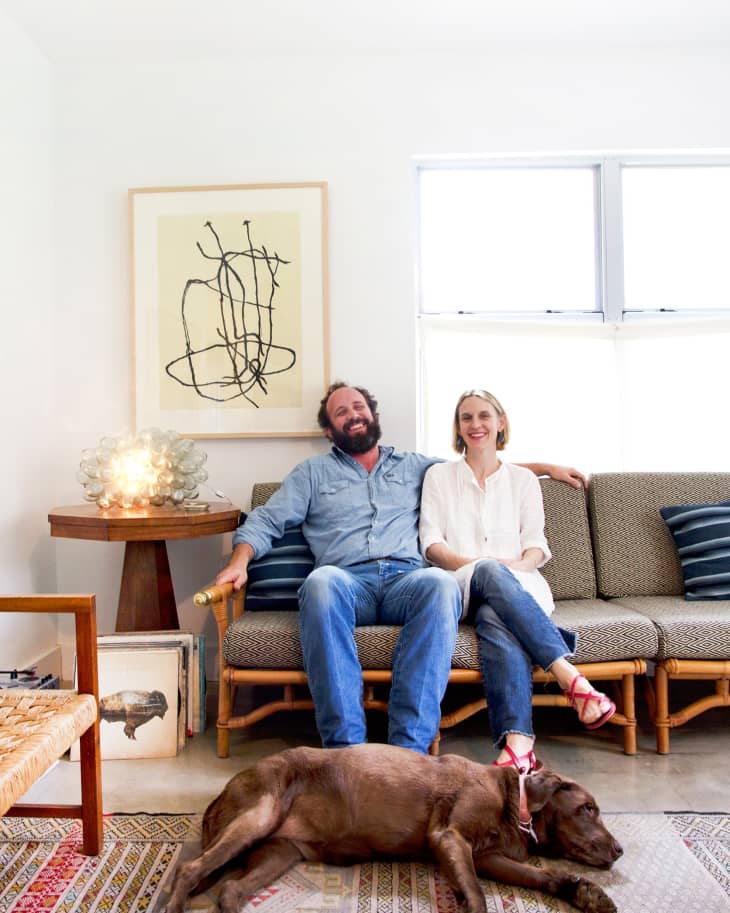 Couple sitting on a sofa with a brown dog lying on a patterned rug, abstract art on the wall, and a side table with a lamp.