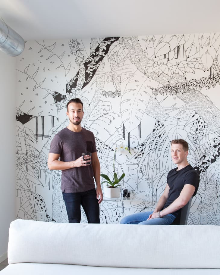 Two men in a room with a black and white botanical mural, one standing with a drink, the other seated near a small table with an orchid.