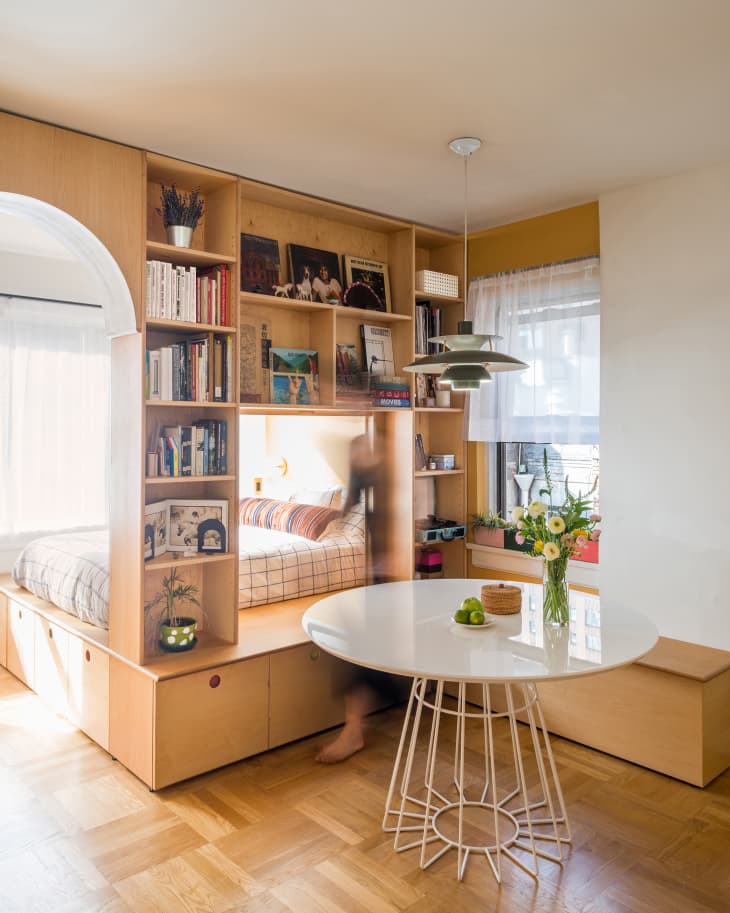 Built in wood cabinets, including a bookshelf and bench, separating a bedroom from a dining room