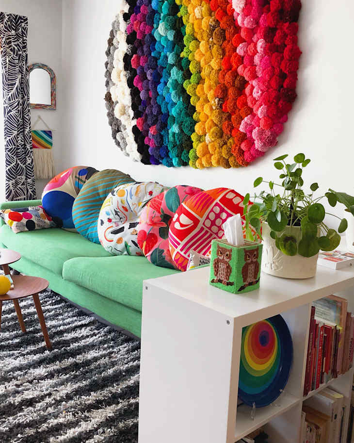 Colorful living room with a green sofa, vibrant round cushions, rainbow wall art, and a plant on a white shelf.