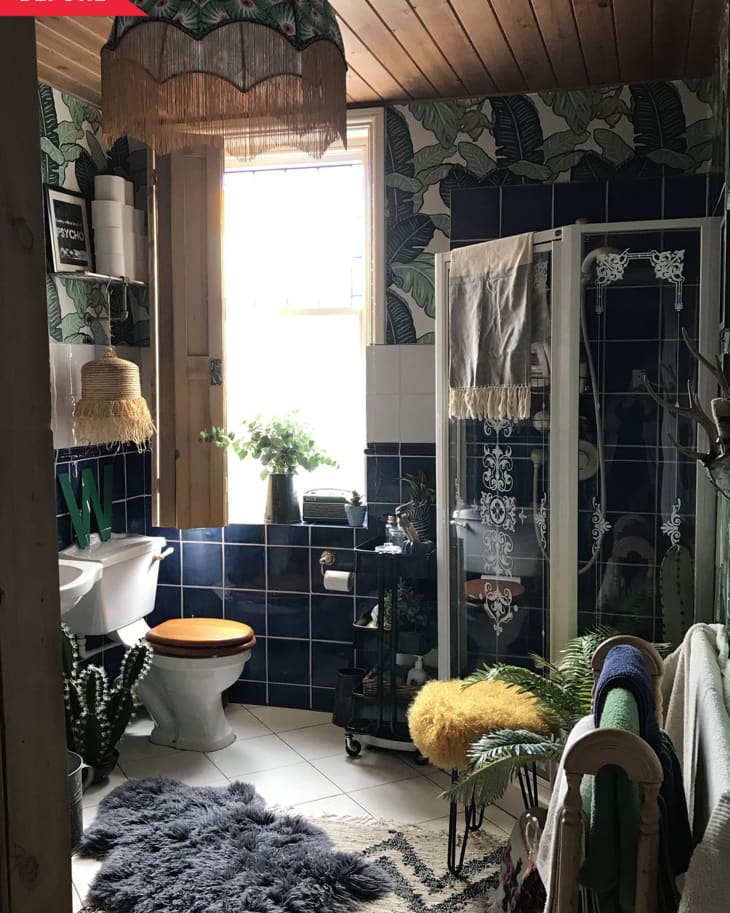 Bathroom with dark blue tiles, leafy wallpaper, wooden ceiling, and eclectic decor including a fringed lamp and fluffy rug.