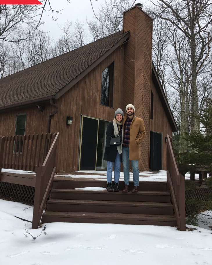 Couple standing on snowy steps in front of a wooden A-frame cabin with large windows and a tall chimney.