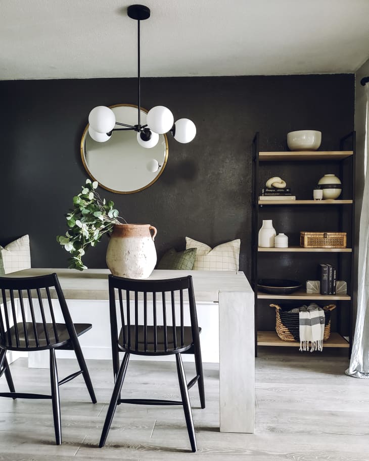 Dining area with black chairs, white table, round mirror, and modern chandelier. Shelves with decor items and a woven basket.