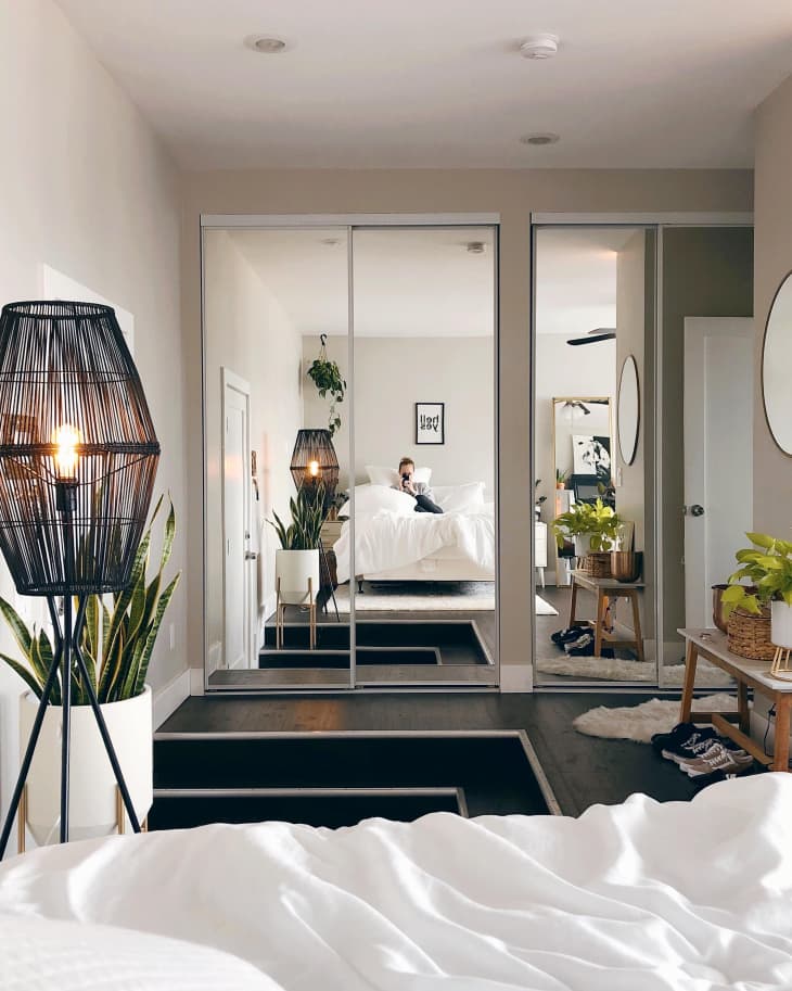 Modern bedroom with mirrored closet doors, black floor lamp, plants, and a person sitting on a white bed.