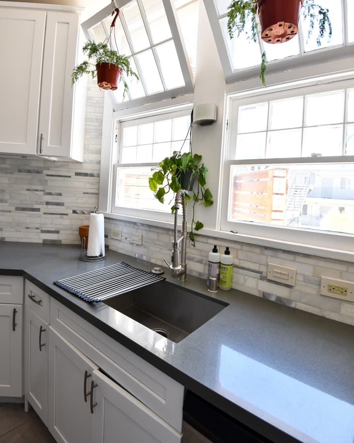 Kitchen with large paned open windows, hanging plants, stone tile backsplash, gray countertops, white cabinets
