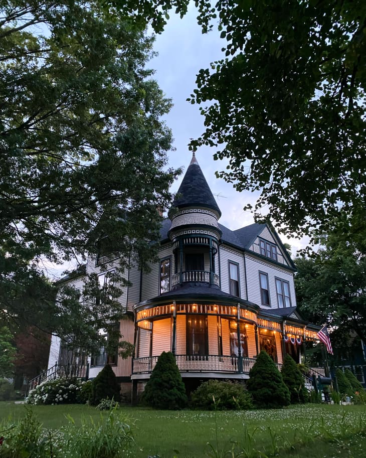 Victorian-style house with a turret, wraparound porch, and warm lights, surrounded by trees and a well-kept lawn.
