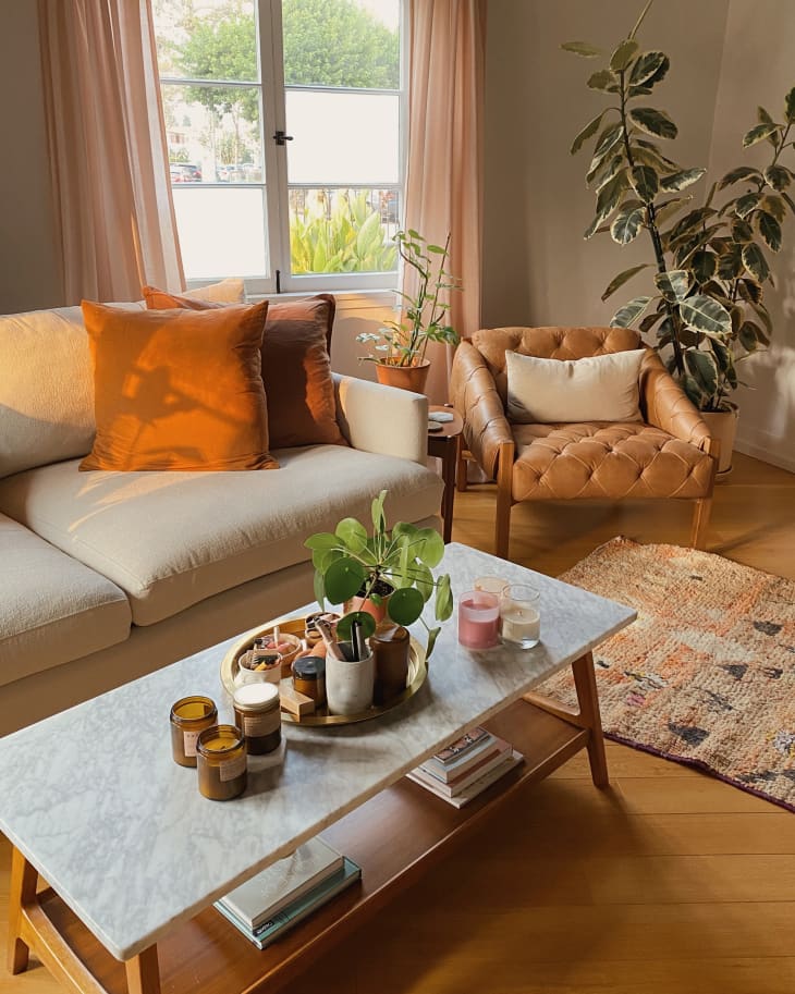 Cozy living room with a cream sofa, orange pillows, marble coffee table, candles, and potted plants by a window.