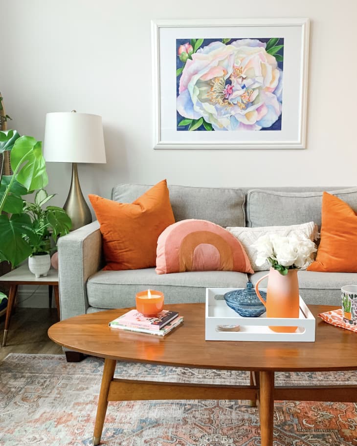 Gray sofa with orange pillows, wooden coffee table, plants, and floral artwork in a cozy living room.