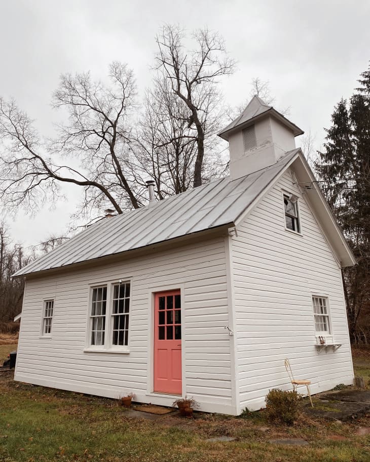 White wooden cottage with a pink door, metal roof, and small bell tower, surrounded by bare trees.