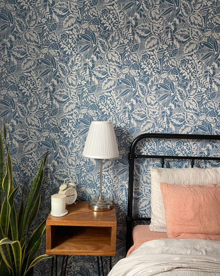 Bedroom with floral wallpaper, black metal bed, pink pillows, wooden nightstand, lamp, clock, candle, and a potted plant.