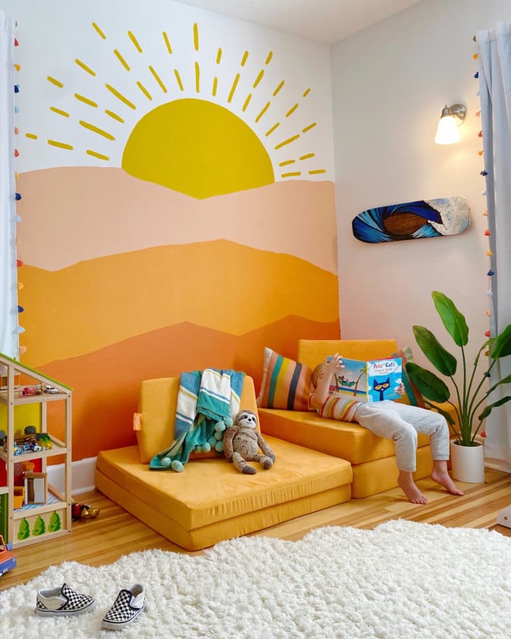 Child reading on yellow cushions in a colorful playroom with a sun mural, toy shelf, and plant.