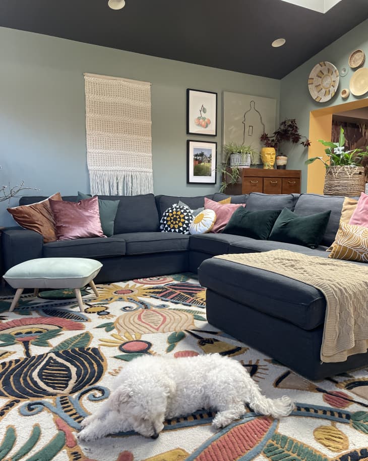 A living room with green walls, a large gray sectional covered in colorful pillows, and a floral rug.