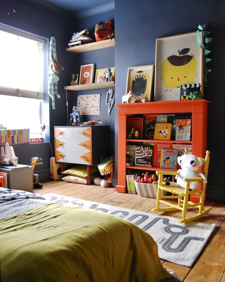 Children's room with blue walls, orange bookshelf, yellow rocking chair, and colorful decor.