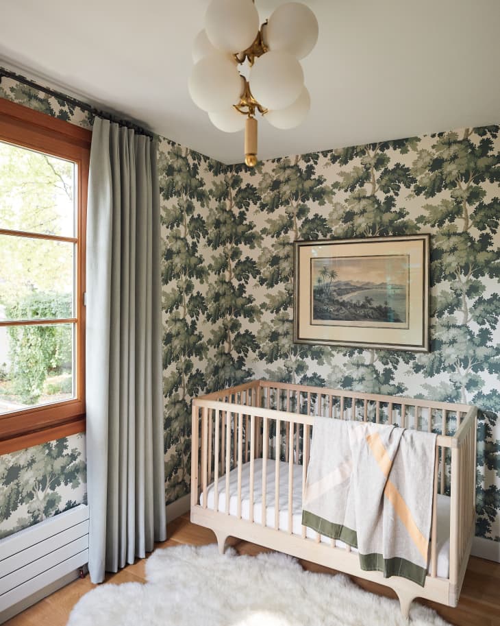 Nursery with a wooden crib, leafy wallpaper, light curtains, and a modern chandelier.